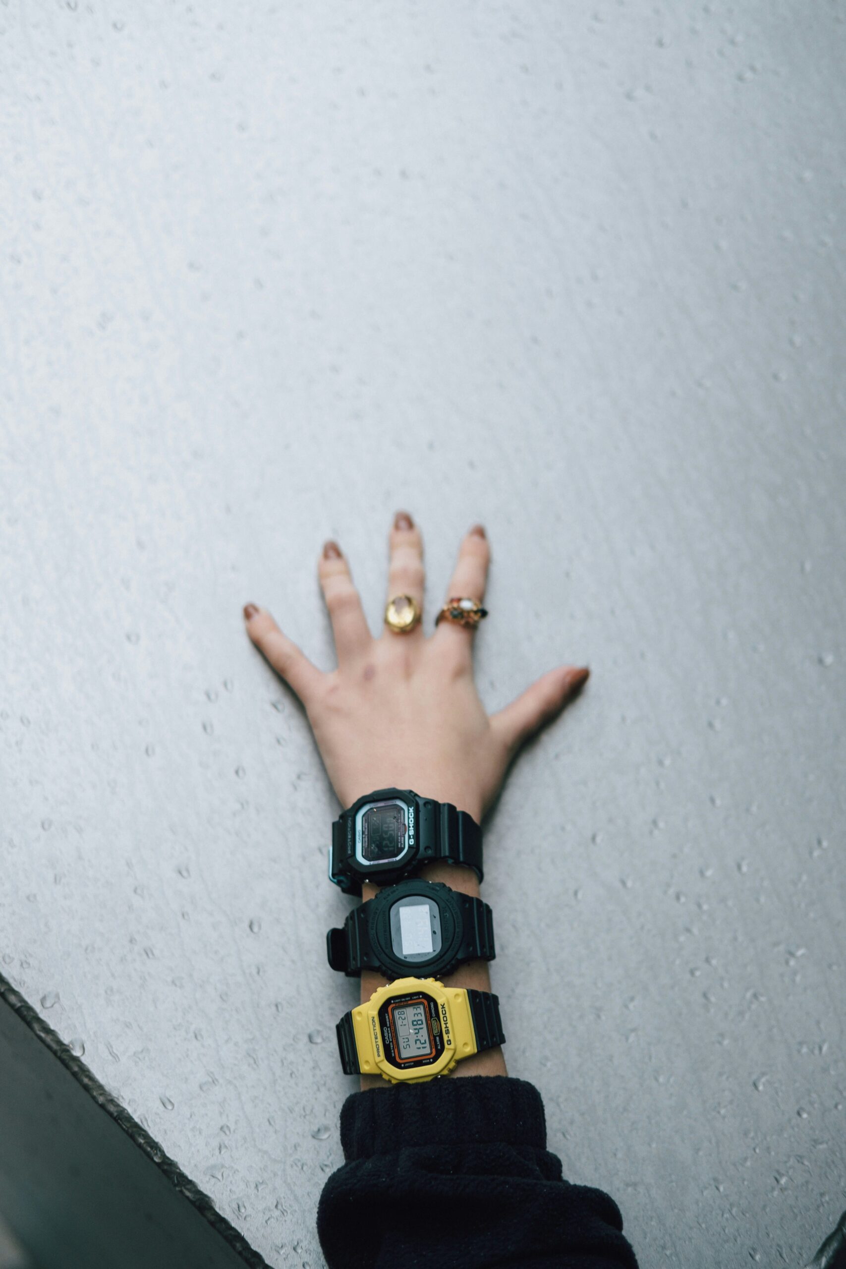 Mano con tres relojes digitales de colores verde, negro y amarillo, apoyada en una pared gris.