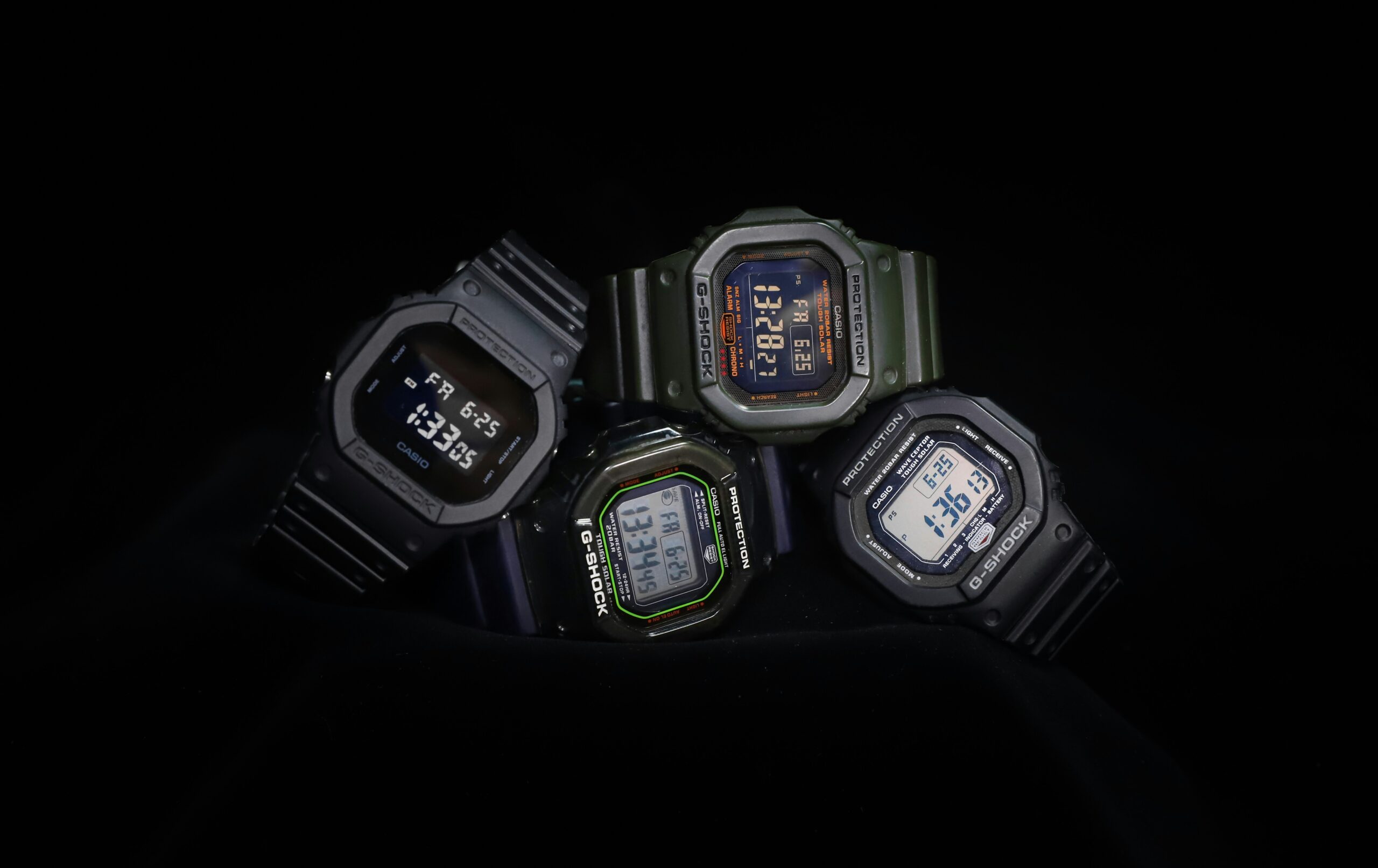 Cuatro relojes G-Shock en tonos negro y verde militar sobre fondo oscuro, mostrando diferentes diseños digitales.