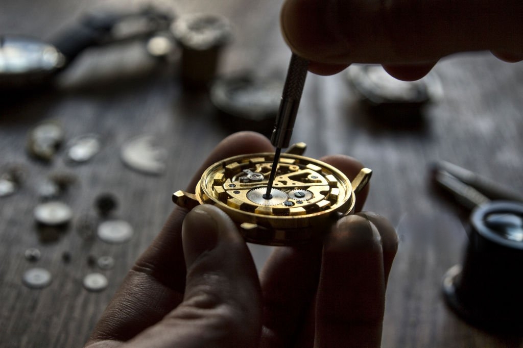 Manos de relojero ajustando un mecanismo de reloj dorado con herramientas sobre una mesa de trabajo.